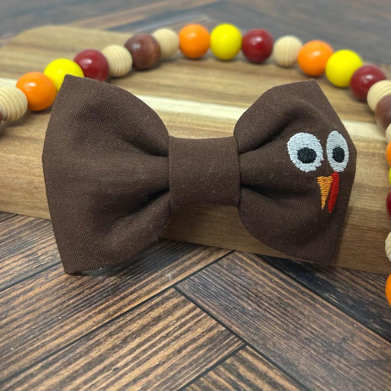 May include: A brown bow tie with a turkey face embroidered on the right side. The bow tie is on a wooden surface with a beaded necklace in the background. The beads are in shades of orange, yellow, red, and brown.