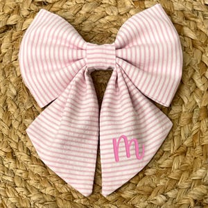 May include: A pink and white striped fabric bow tie with the letter "M" embroidered in pink.