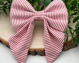 May include: A fabric hair bow featuring a pattern of thin, vertical red and white stripes. The bow is constructed with two large, folded loops and two long, pointed tails extending downward, creating a symmetrical, multi-layered appearance.