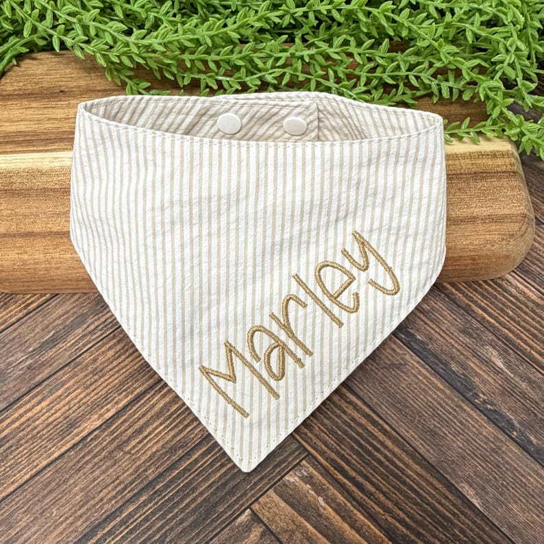 May include: A beige and white striped dog bandana with the name "Marley" embroidered in a gold color. The bandana has two white snap closures at the top and is triangular in shape. The background is a wooden surface with greenery.
