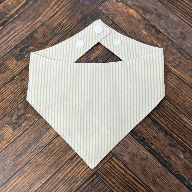 May include: Cream and white striped bandana bib with two white snap closures. The bib is made of soft fabric and is designed to catch drool or spills. The bib is a stylish and practical accessory for babies and toddlers.