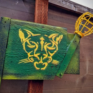 May include: A wooden sign with a yellow cougar silhouette painted on a green background. A yellow metal cage light fixture is attached to the sign.