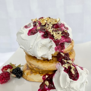 May include: A stack of three golden brown pancakes topped with whipped cream, a red berry sauce, and edible gold flakes.