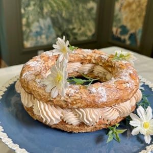 Puede incluir: Un postre de pastelería en forma de anillo, con un exterior marrón dorado espolvoreado con azúcar glas y adornado con flores blancas. El postre se encuentra sobre un plato azul con un borde decorativo blanco.