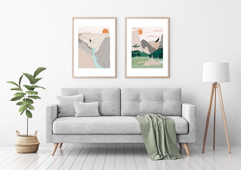 May include: Two framed illustrations of mountain landscapes with a sunset in the background. The illustrations are in a minimalist style with muted colors and simple lines. The illustrations are hung on a white wall above a gray sofa.