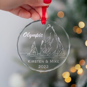 May include: A clear glass Christmas ornament with a white etched design of two mountains and the text "Olympic" above the mountains. The ornament also has the text "Kirsten & Mike 2022" below the mountains.