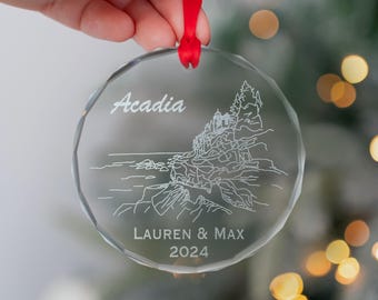 May include: A clear glass ornament with an etched design of a coastal scene with a lighthouse and the words "Acadia" and "Lauren & Max 2024".