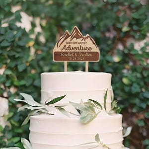 May include: A wooden cake topper featuring a mountain range design with the words "OUR GREATEST Adventure" and the names "Rachel & Stephen" with the date "10.24.2025". The topper is placed on a white tiered cake, decorated with greenery.