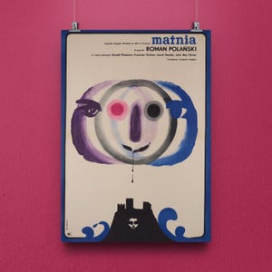 May include: A framed vintage movie poster with a blue border. The poster features an abstract face with purple, pink, gray, and blue elements. Below the face is a black silhouette of a castle and waves. The Polish title "Matnia" is at the top.