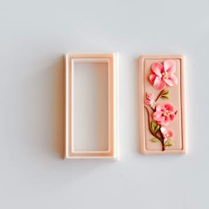 May include: A pink and green floral design cookie cutter with a rectangular frame. The cutter is made of plastic and has a smooth surface.