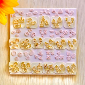 May include: A selection of light pink and yellow cookie cutters in various shapes, including seashells, fish, and palm trees, arranged on a white surface. A yellow sunflower is in the upper left corner.
