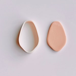 May include: A white plastic cookie cutter shaped like an egg next to a light peach-colored piece of clay.