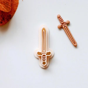 May include: Two plastic cookie cutters in the shape of a sword. The cutters are white and have a detailed design.