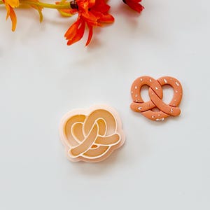 Puede incluir: Dos abalorios de arcilla polimérica con forma de pretzel. Uno es de color melocotón claro con un contorno blanco, y el otro es de color melocotón más oscuro con detalles blancos. Flores naranjas y amarillas en el fondo.