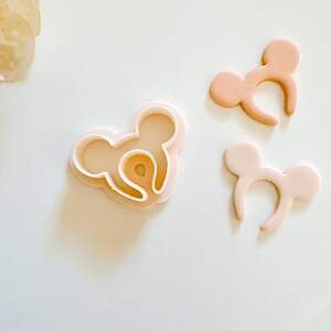May include: A set of three pink plastic cookie cutters shaped like Mickey Mouse ears. The cutters are used to create cookies or fondant decorations.