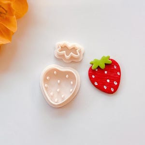 Puede incluir: Un juego de manualidades con temática de fresas que presenta una fresa roja, un cortador de galletas con forma de fresa y un cortador con forma de hoja. La fresa tiene puntos blancos y una parte superior verde. Los cortadores son de color rosa claro.