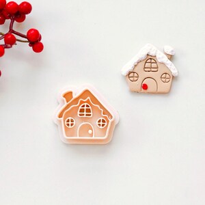 May include: Two gingerbread house cookie cutters, one white and one pink, on a white background. The cutters are shaped like houses with windows and doors.