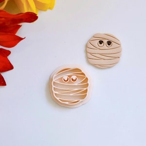 May include: Two beige plastic cookie cutters shaped like mummies with eyes. The cutters are on a white background.