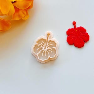 Puede incluir: Un cortador de galletas de plástico blanco con forma de flor de hibisco. El cortador se muestra junto a una flor de hibisco roja.