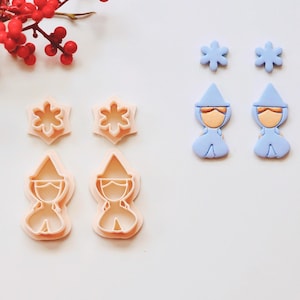 May include: Two sets of cookie cutters, one set of two snowflake cutters and one set of two gnome cutters. The cutters are pink and the gnomes have a white hat and a blue body. There are also two blue gnome-shaped polymer clay earrings.
