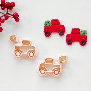 May include: A selection of red truck-shaped cookie cutters and two red truck-shaped biscuits with green tree details. The cookie cutters are light orange and have a truck outline. The biscuits have black wheels. A sprig of red berries is in the upper left corner.