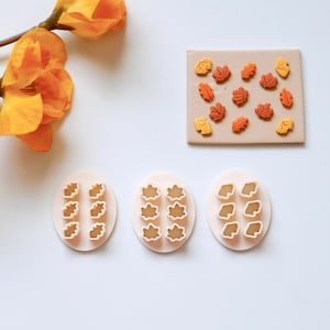 Puede incluir: Tres cortadores de galletas de plástico blanco con diseños de hojas. Los cortadores están sobre una superficie blanca. También hay varias galletas pequeñas en forma de hojas de color naranja y amarillo sobre una superficie marrón.