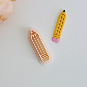 May include: Two pencil-shaped cookie cutters, one yellow and one white. The yellow cutter has a pink eraser and the white cutter is a mold.