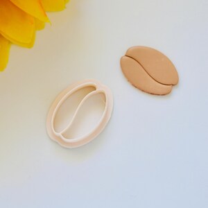 May include: A white plastic cookie cutter shaped like a coffee bean, next to a brown coffee bean cookie.