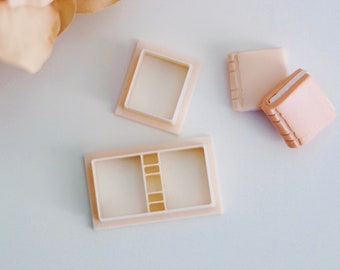 May include: A set of three white plastic molds for making miniature books. The molds are in the shape of open books with a rectangular shape and a slightly raised spine. The molds are on a white surface.