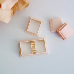 May include: A set of three white plastic molds for making miniature books. The molds are in the shape of open books with a rectangular shape and a slightly raised spine. The molds are on a white surface.
