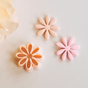 May include: Three pink plastic flower-shaped cookie cutters. Two of the cutters are solid and one is open. The open cutter is slightly larger than the solid cutters.