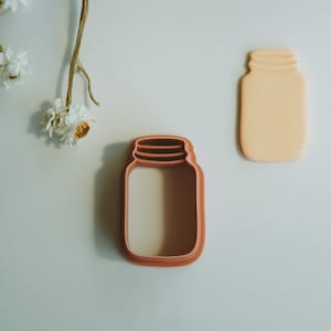 May include: A brown plastic cookie cutter shaped like a mason jar with a lid. The cookie cutter is on a white surface.