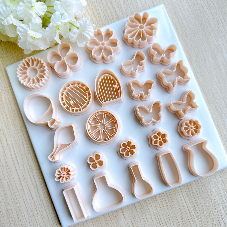 Pu&ograve; includere: Un set di 20 stampini per biscotti in varie forme, tra cui fiori, farfalle, una ruota di carro, una casa e un vaso. Gli stampini sono in plastica rosa chiaro e sono disposti su una superficie bianca.