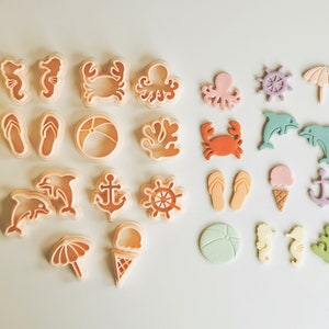 May include: A set of 18 plastic cookie cutters in various summer and beach themes. The cutters are in shades of pink, blue, orange, and white. The cutters include a crab, starfish, seashell, flip-flop, anchor, ice cream cone, umbrella, and more.