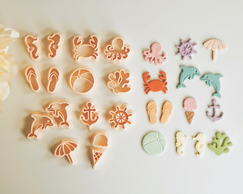 May include: A set of 18 plastic cookie cutters in various summer and beach themes. The cutters are in shades of pink, blue, orange, and white. The cutters include a crab, starfish, seashell, flip-flop, anchor, ice cream cone, umbrella, and more.