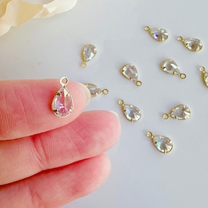 May include: A close-up of a hand holding a clear teardrop-shaped crystal pendant with a gold bail. The pendant is surrounded by other similar pendants on a white surface.