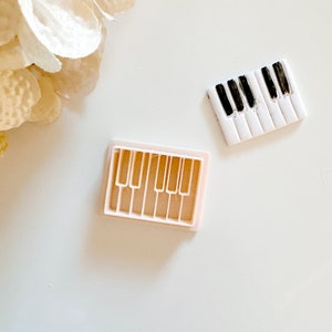 May include: Two white plastic molds for making fondant or cookie decorations. One mold is shaped like a piano keyboard with black keys. The other mold is shaped like a piano keyboard with seven keys.