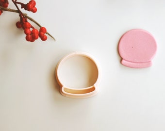 May include: Two cookie cutters, one in the shape of a snow globe and one in the shape of a solid circle. The snow globe cutter is white and the solid circle cutter is pink.