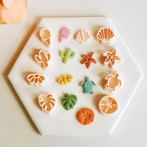 May include: A set of 14 plastic cookie cutters in various shapes, including a seashell, a starfish, a turtle, a palm tree, a cactus, an ice cream cone, and a flower. The cutters are arranged on a white hexagonal surface.