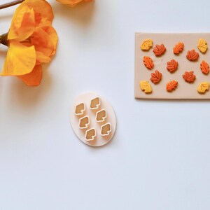 Puede incluir: Un molde de plástico blanco con seis recortes en forma de hoja. Junto a él hay una lámina de arcilla marrón con varias formas de hojas naranja y marrón cortadas.