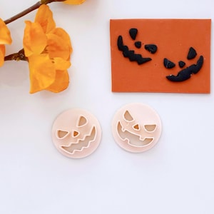 May include: Two pink plastic pumpkin cookie cutters with a scary face design. The cutters are on a white surface.