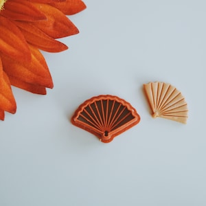 May include: Two red and brown plastic cookie cutters shaped like fans. The fan on the left is a darker red and the fan on the right is a lighter brown.