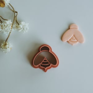 May include: Two bee-shaped cookie cutters, one pink and one brown. The pink cutter is on the right and the brown cutter is on the left.