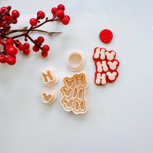 May include: A set of white plastic cookie cutters in the shape of the letters "HO" and a Mickey Mouse head. The cutters are on a white surface with a red berry branch and a red circle in the background.