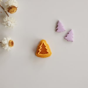 May include: A yellow plastic Christmas tree cookie cutter with a brown caramel-colored tree inside. Two purple plastic Christmas tree cookie cutters are in the background.