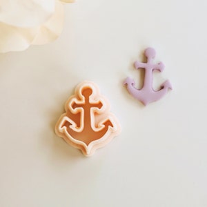 May include: Two anchor cookie cutters, one is a light pink color and the other is a light peach color. The cutters are on a white surface.