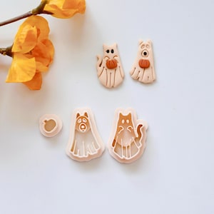 May include: Set of five cookie cutters in the shape of ghosts and pumpkins. The ghosts have cat and dog faces and are holding pumpkins. The cutters are made of a light pink plastic.