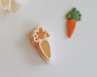May include: A carrot-shaped cookie cutter and a small carrot-shaped decoration. The cookie cutter is light orange and has a layered design. The carrot decoration is orange with green leaves. The items are on a white surface.