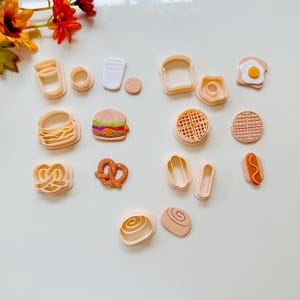 Peut inclure: Ensemble de découpoirs à biscuits sur le thème de la nourriture, comprenant un hamburger, un bretzel, une gaufre et un hot-dog. Les découpoirs sont dans des tons beige et marron, avec des détails colorés. L'ensemble est disposé sur une surface blanche.