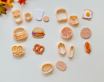 Könnte beinhalten: Eine Sammlung von Ausstechern in Form von Lebensmitteln, darunter ein Hamburger, eine Brezel, eine Waffel und ein Hotdog. Die Ausstecher sind in Beige- und Brauntönen gehalten, einige mit farbigen Details. Die Anordnung befindet sich auf einer weißen Oberfläche.